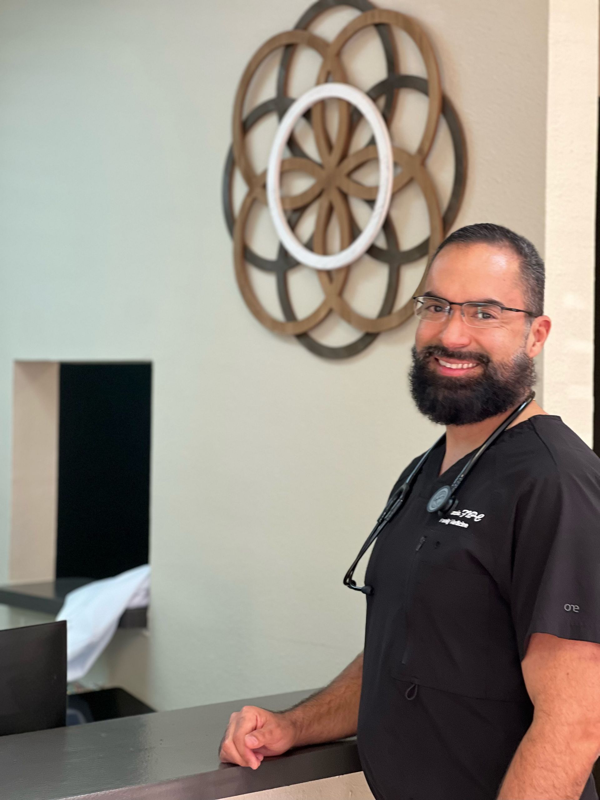 Smiling man in black scrubs standing at a clinic reception desk near a wall sculpture.