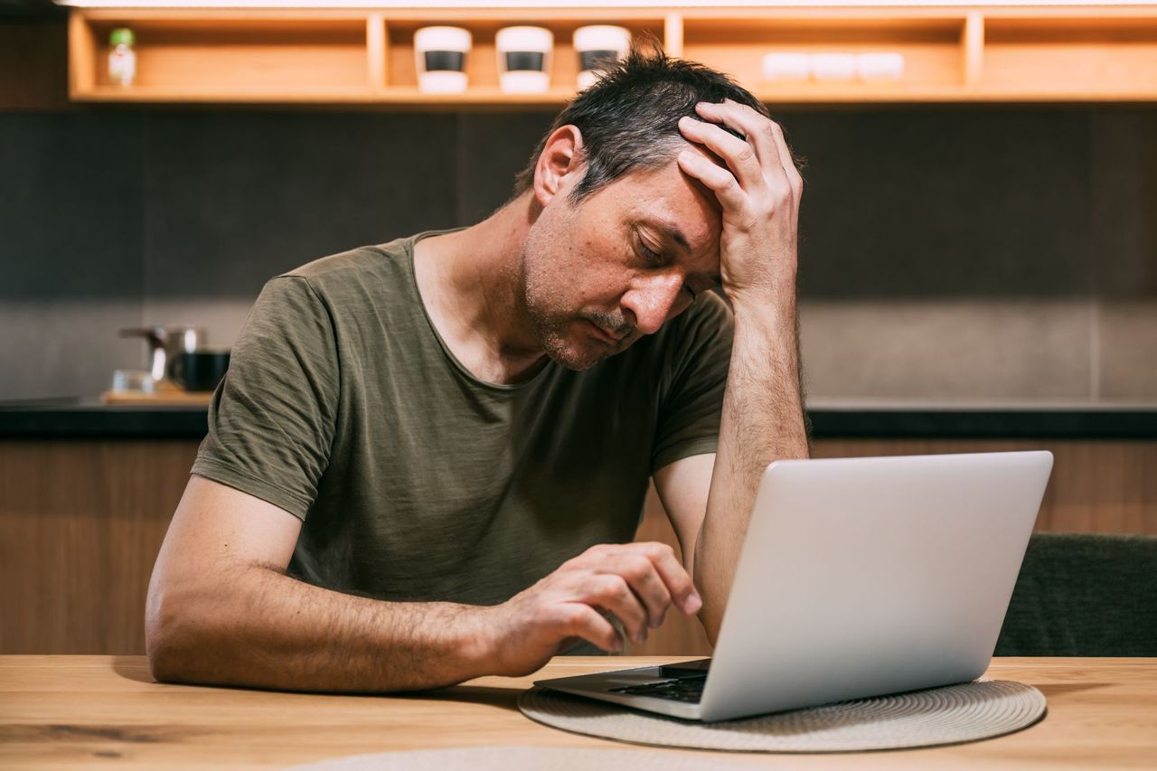Man sitting at a table, looking stressed while using a laptop in a modern kitchen