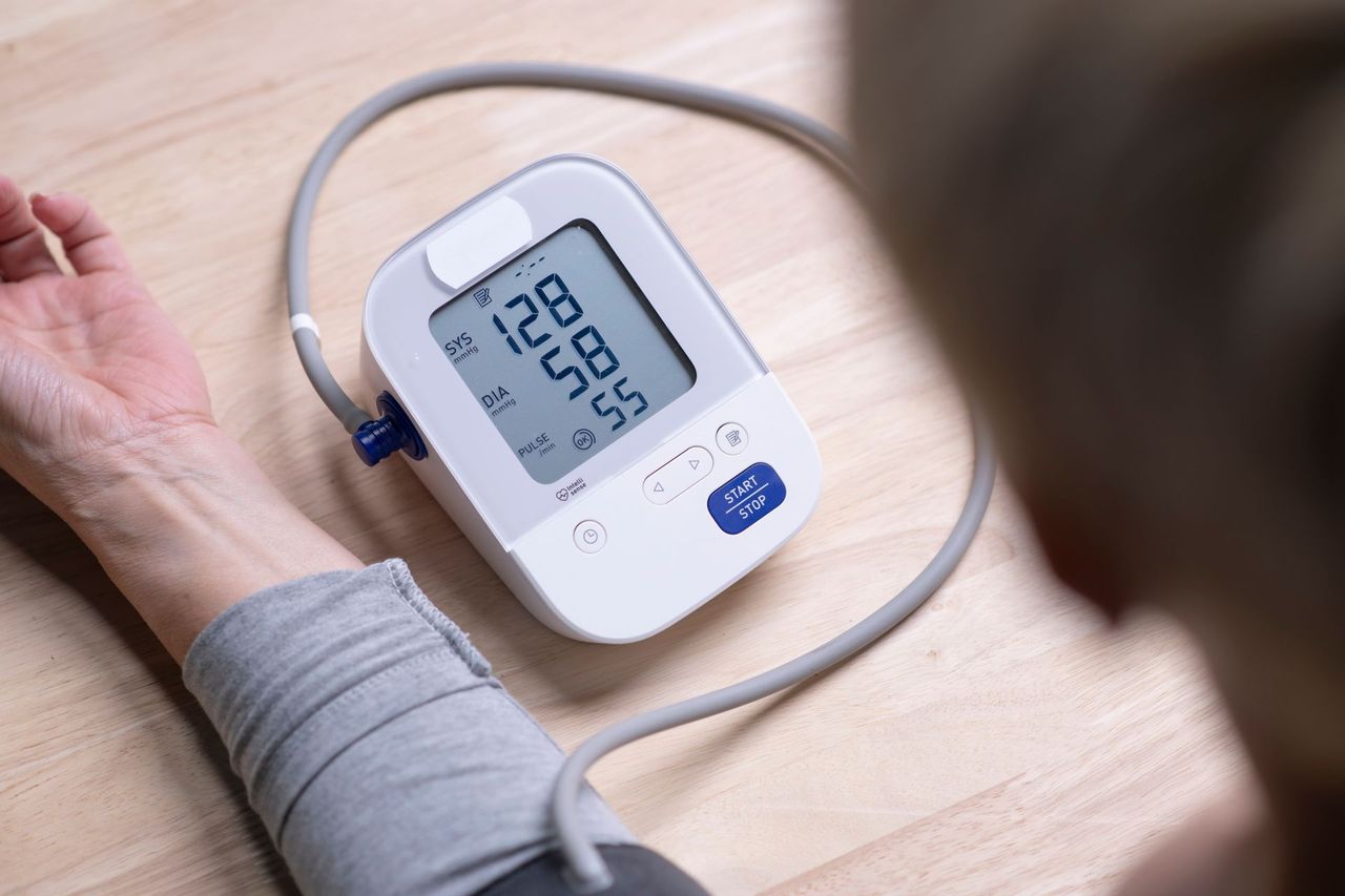 Arm with blood pressure cuff on wooden table, digital monitor showing 128/58 and pulse 56.