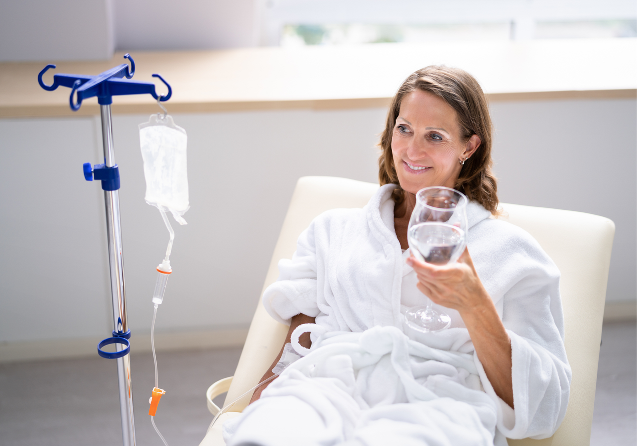 Woman in a hospital chair holding a glass of water beside an IV stand