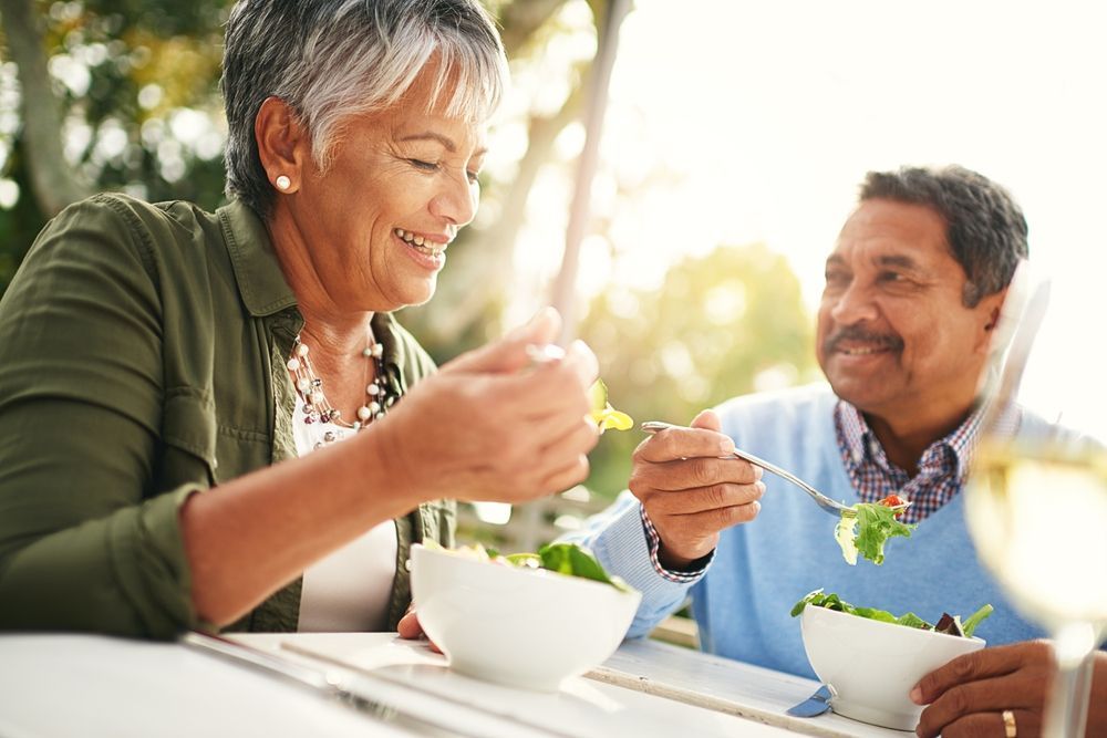 Two people smiling and eating together outdoors at a table with bowls in warm sunlight