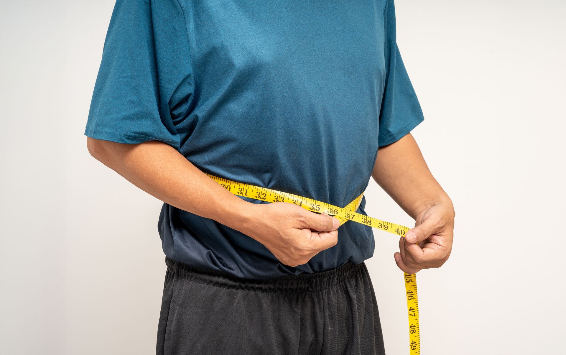 Person measuring waist with a yellow tape measure against a blue shirt and dark pants