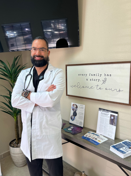Bearded doctor in white coat standing in a clinic lobby beside a framed quote and brochures.